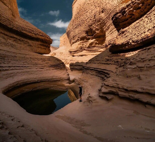 A person standing in a narrow canyon with layered rock formations and a small pool of water. 1