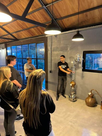 A group of people observing a distillation apparatus in a room with wooden beams and large windows. 2