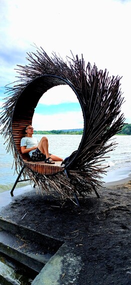A person sitting on a circular, straw-wrapped chair by a lake. 1