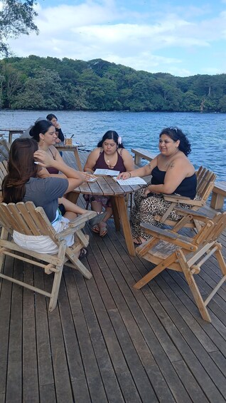 A group of people seated at a wooden table on a dock by a lake, with chairs arranged around the table. 3