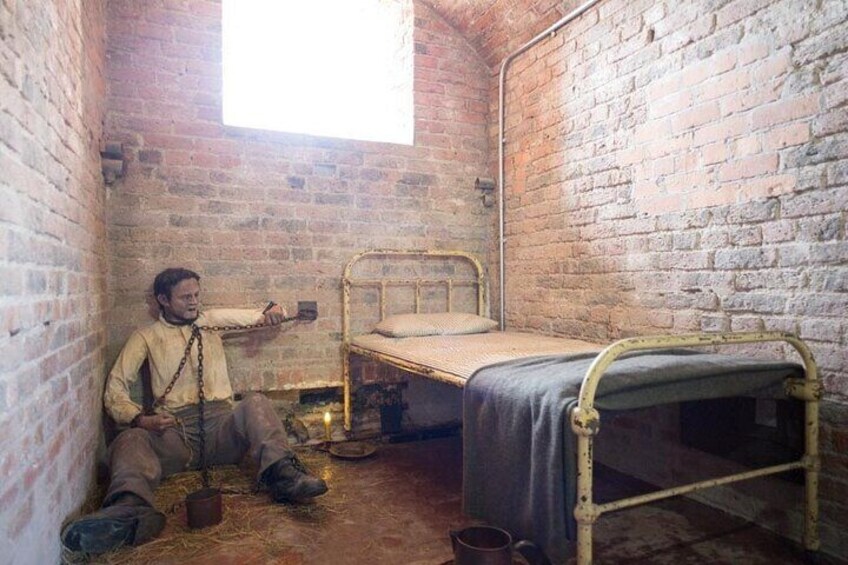 A brick-walled room with a bed and a person chained to a wall. 1
