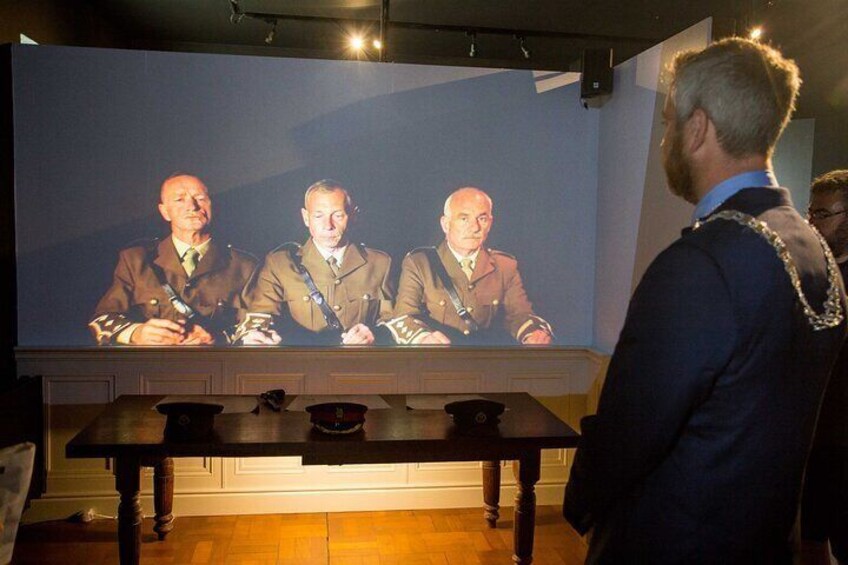 A person standing in front of a large screen displaying three men in suits. 5