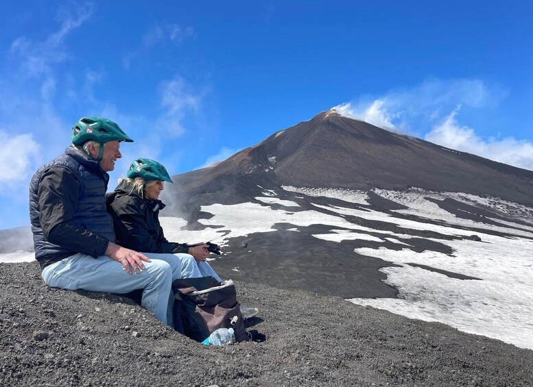 Two people sitting on a rocky surface with a snow-covered mountain and a clear sky in the background. 4