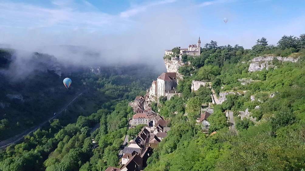 A mountainous landscape with a historic castle perched on a cliff, hot air balloons in the sky, and a dense forested valley below. 1