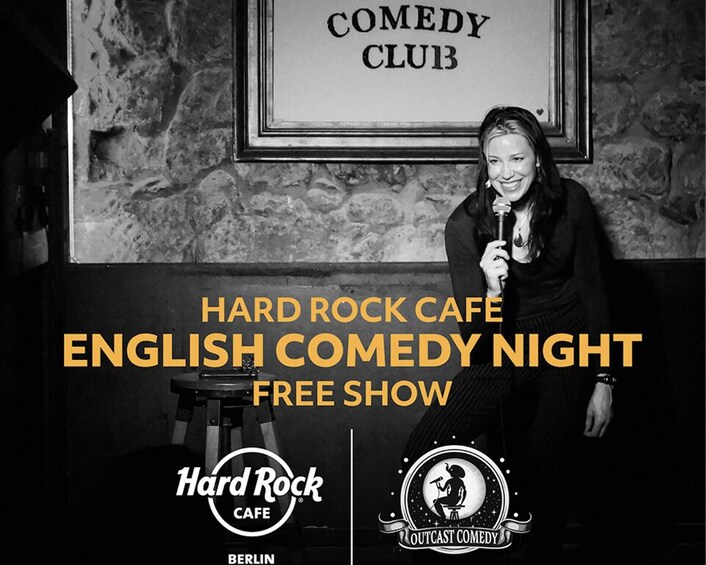 A person holding a microphone in front of a sign that reads 'COMEDY CLUB' in a venue with a stone wall background. 1