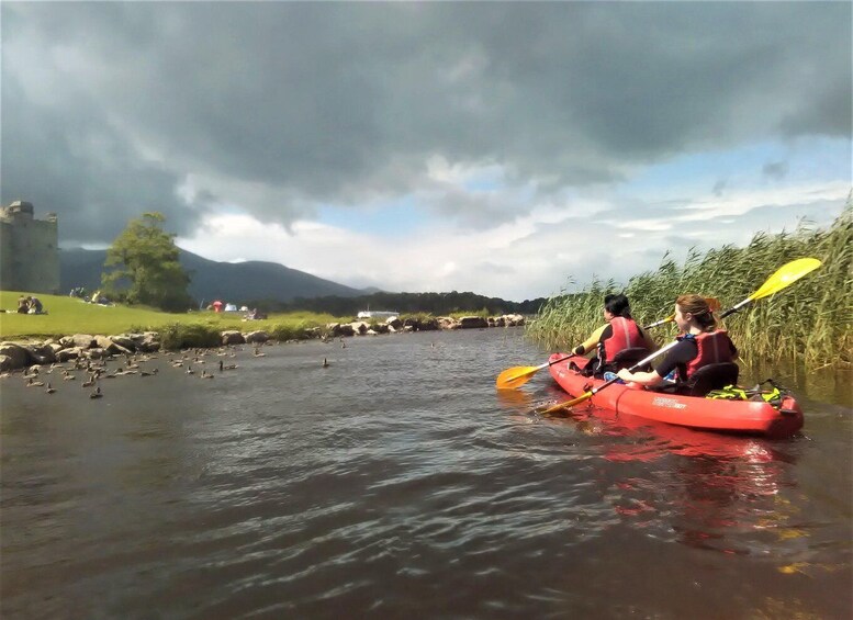 Two people kayaking on a river with ducks, a castle in the background, and a cloudy sky. 1