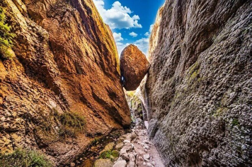 A narrow canyon with a rock formation balancing on a ledge, a rocky trail, and a clear blue sky. 4