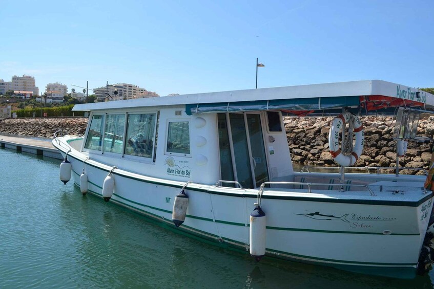 A white boat named 'Alvor Flor do Sol' docked at a marina with a rocky shoreline and buildings in the background. 2