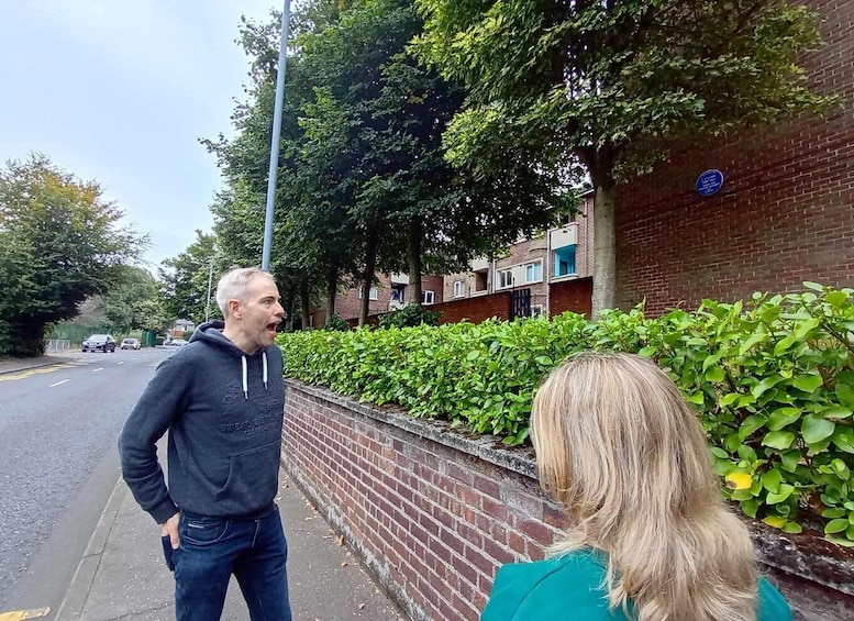 Two people conversing on a sidewalk with a brick wall and greenery in the background. 3