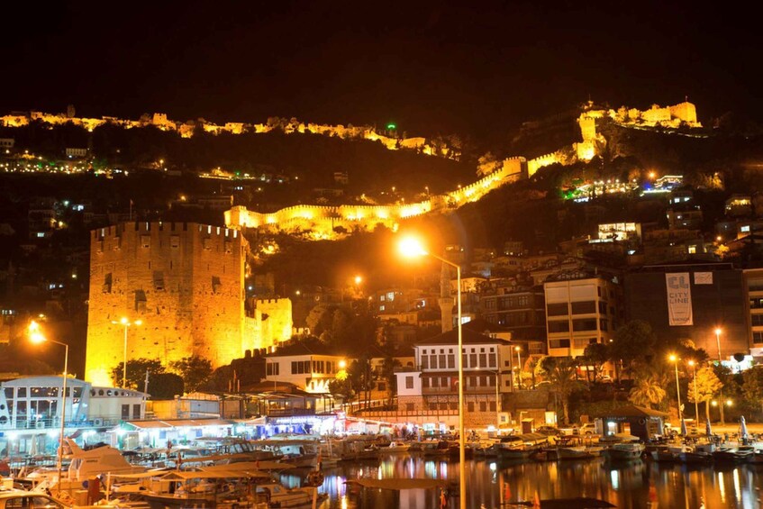 A historic cityscape at night with illuminated fortifications, a prominent castle, and a marina with boats. 2
