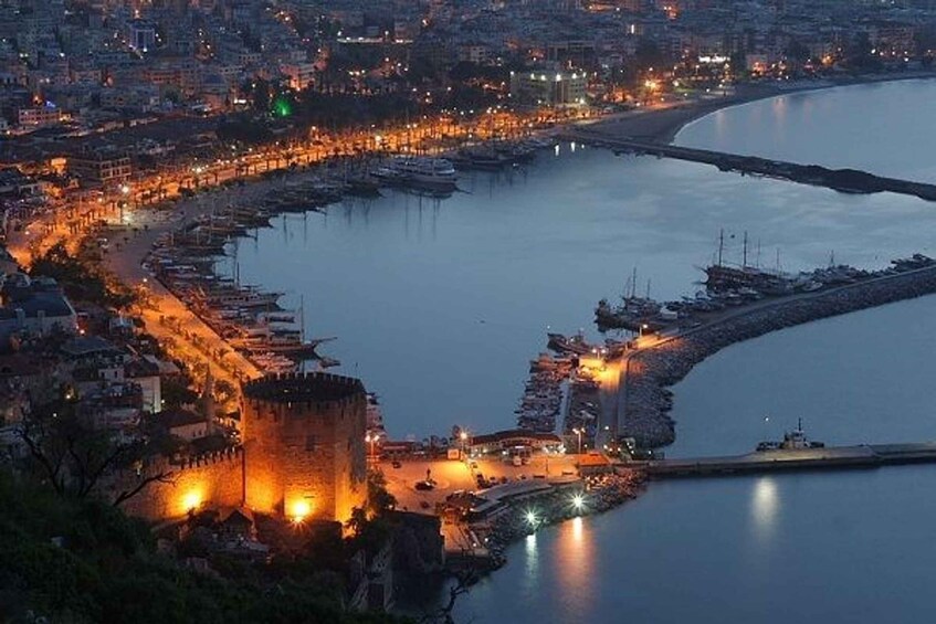 A coastal cityscape at dusk with illuminated buildings, a prominent castle, and a marina with boats. 3