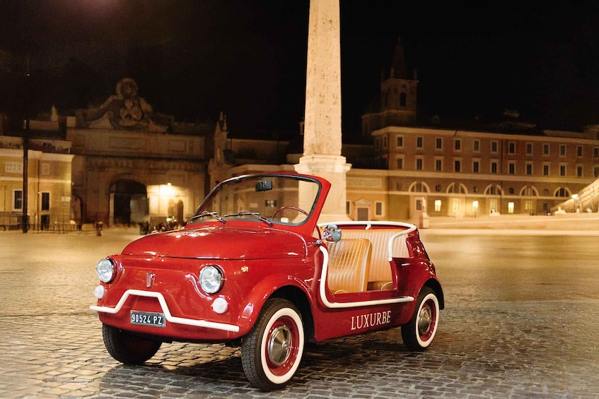 A vintage red car parked on cobblestone pavement with a historic building and a tall monument in the background. 5