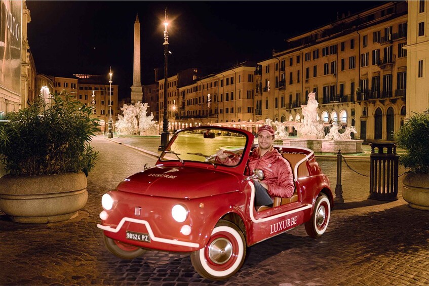 A red vintage car with 'LUXURBE' written on the side parked in front of a historic fountain. 1