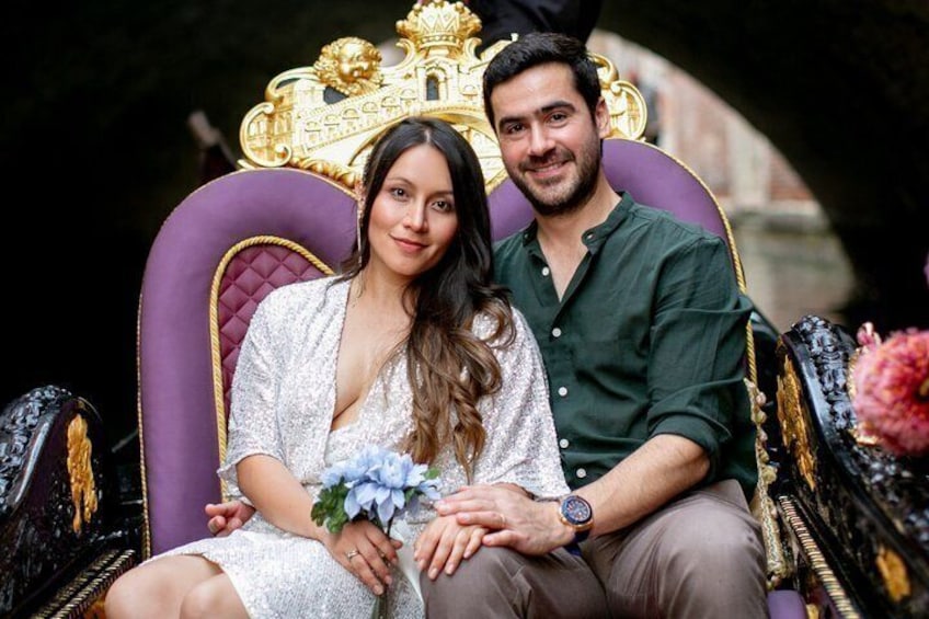 A couple seated on a purple and gold ornate chair with a floral arrangement. 1
