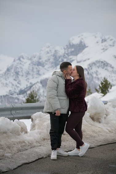 A couple kissing in a snowy mountainous area with snow-covered trees and a guardrail. 2