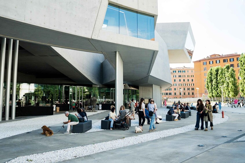 A modern building with a unique geometric design, people sitting and walking in the plaza, and a dog lying on the ground. 5
