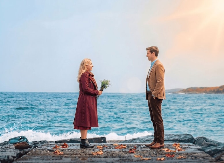 A couple standing on a rocky shore by the sea, with waves crashing in the background. 1