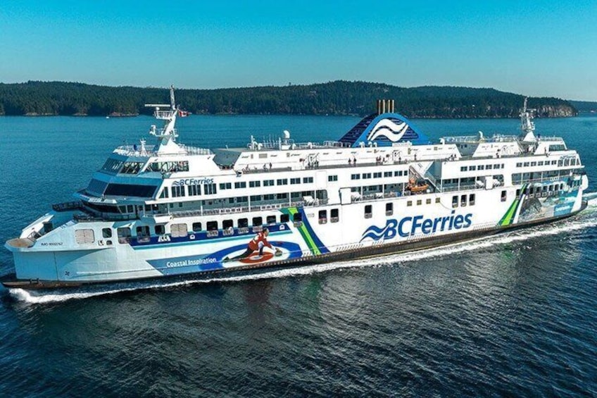 A large ferry with 'BC Ferries' branding, traveling on a body of water with a forested shoreline in the background. 2