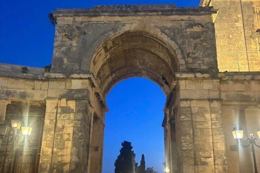 An ancient stone archway with two street lamps on either side, set against a twilight sky. 2