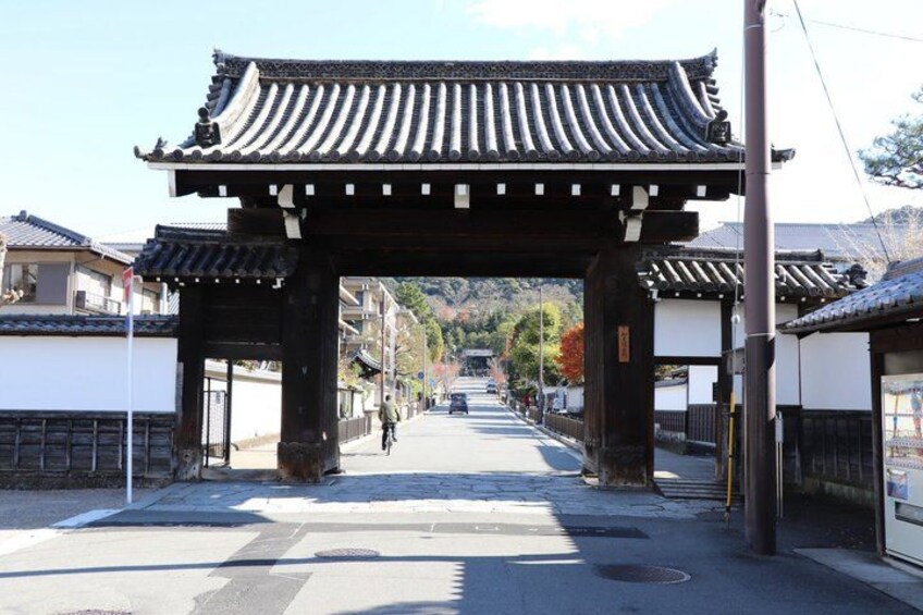 A traditional Japanese gate with a tiled roof, leading to a street lined with buildings and a cyclist in the distance. 2