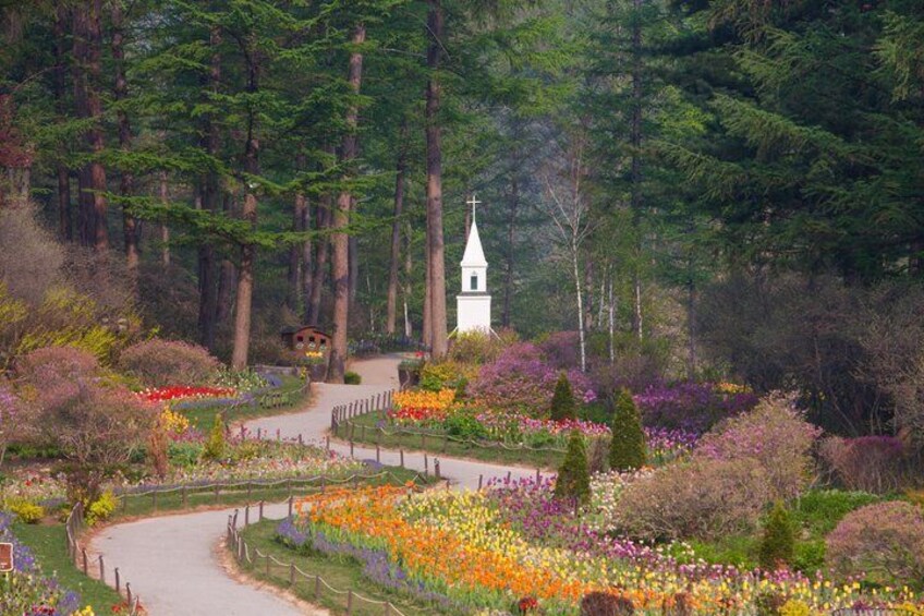 A forested garden with a white church, winding paths, and colorful flower beds. 2