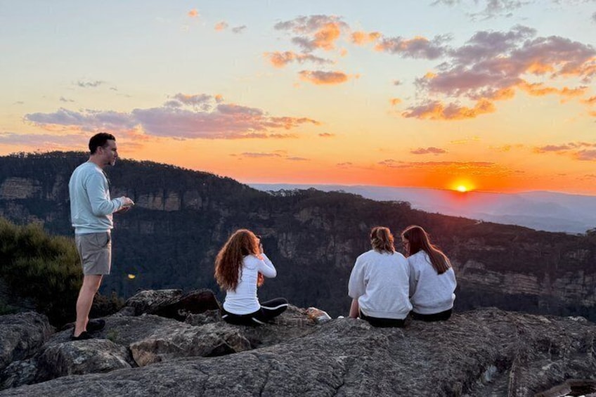 A sunset over a mountainous landscape with three people sitting on rocks. 1