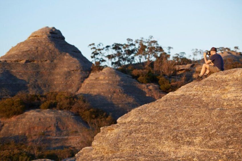 A person sitting on a rock with a camera, surrounded by large boulders and sparse vegetation. 4