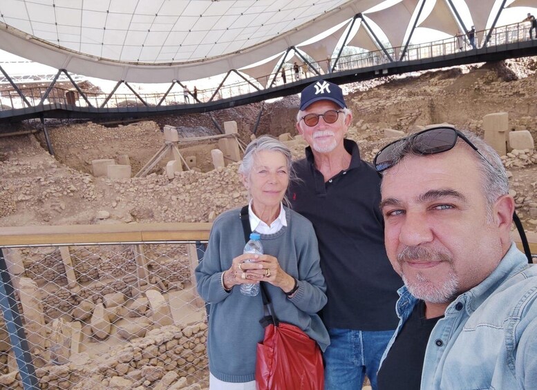 Three people standing in front of an archaeological site with exposed ruins and a protective canopy overhead. 4