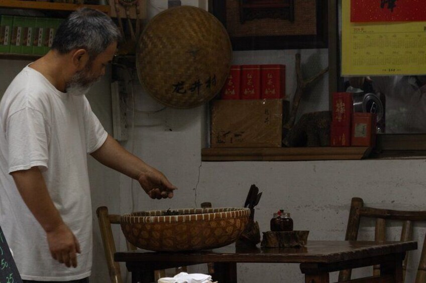 A person in a white shirt standing next to a wooden table with a large woven bowl, a small jar, and red boxes. 3