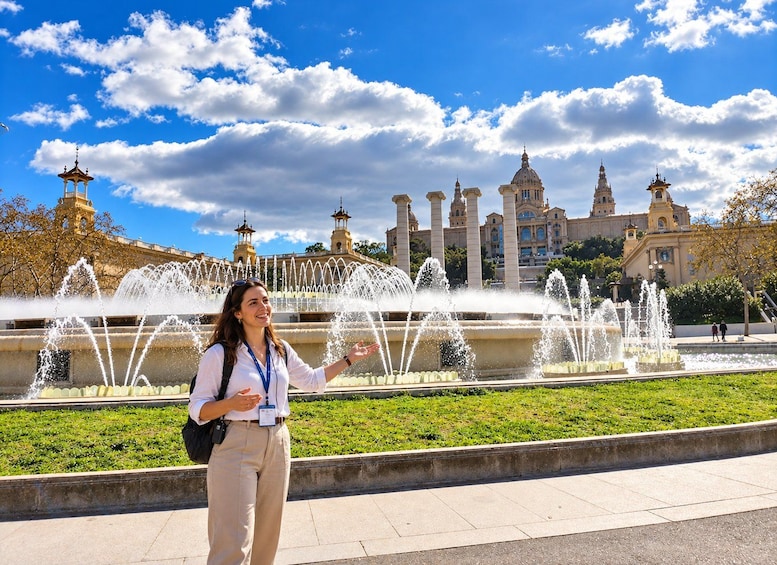 A woman standing in front of a fountain with water jets, with a large building with domes and towers in the background. 1