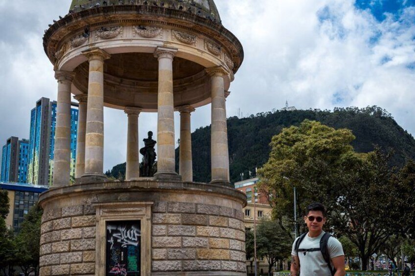 A stone gazebo with columns and a statue, surrounded by trees and buildings, with a hill in the background. 2
