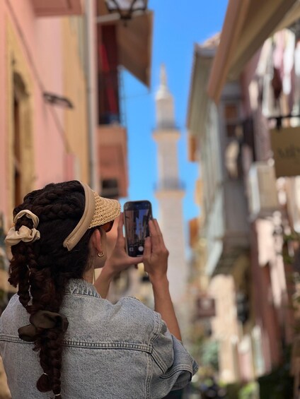 A person taking a photo with a smartphone in a narrow street with buildings and a minaret in the background. 1