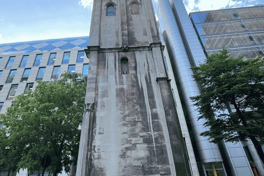 A tall, weathered stone tower with a clock, surrounded by modern buildings and trees. 4