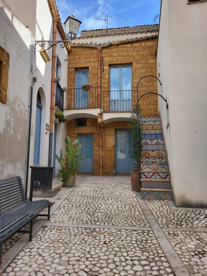 A narrow alley with cobblestone pavement, a bench, a building with a tiled entrance, and a staircase with patterned tiles. 5