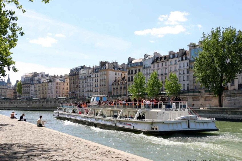 A river cruise boat with passengers, a stone riverbank, and historic buildings along the river. 3