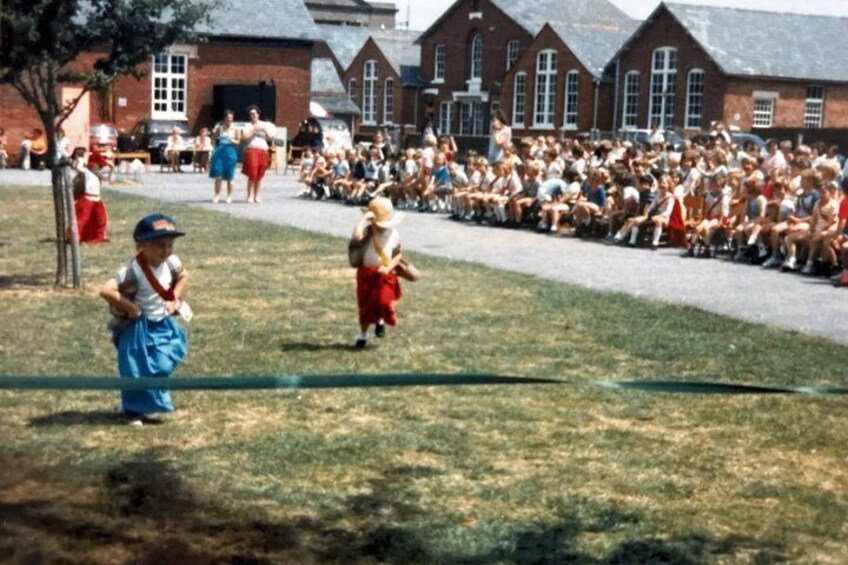 Children in costumes on a school field, spectators in the background, and a brick building. 2