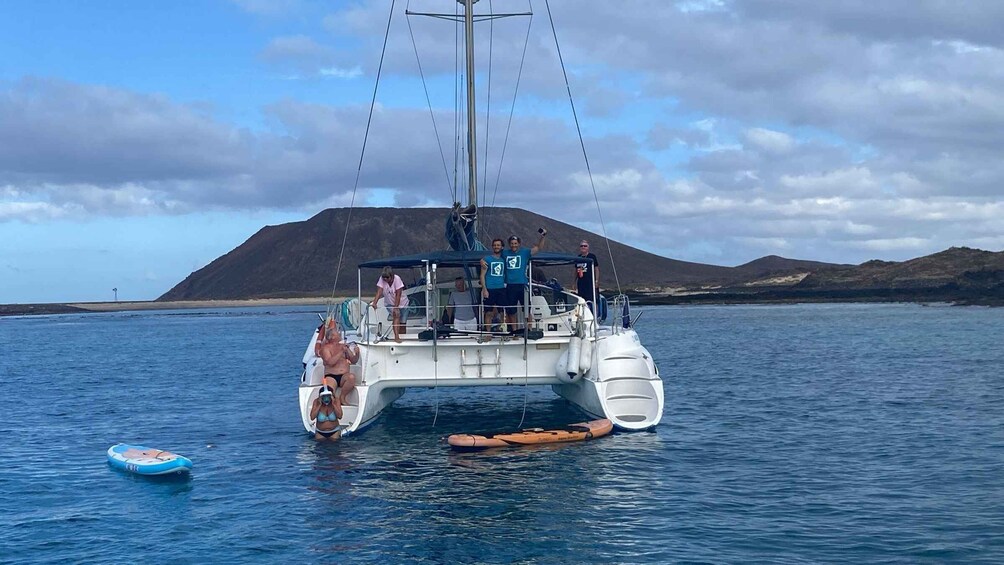 A sailboat with passengers on a calm sea, a small inflatable boat nearby, and a mountainous landscape in the background. 3