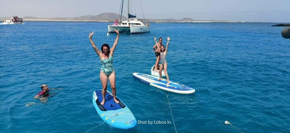 Two people on surfboards in the water with a sailboat and a coastline in the background. 1