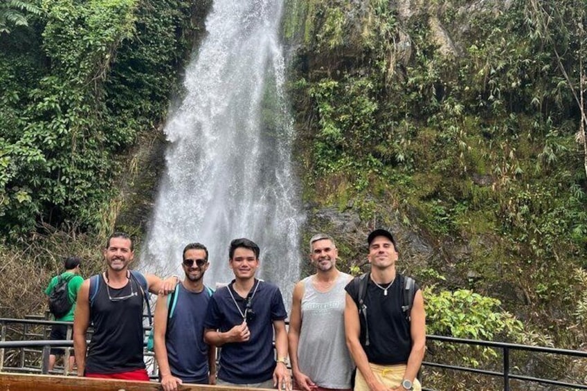 A group of people standing by a waterfall with lush greenery in the background. 1