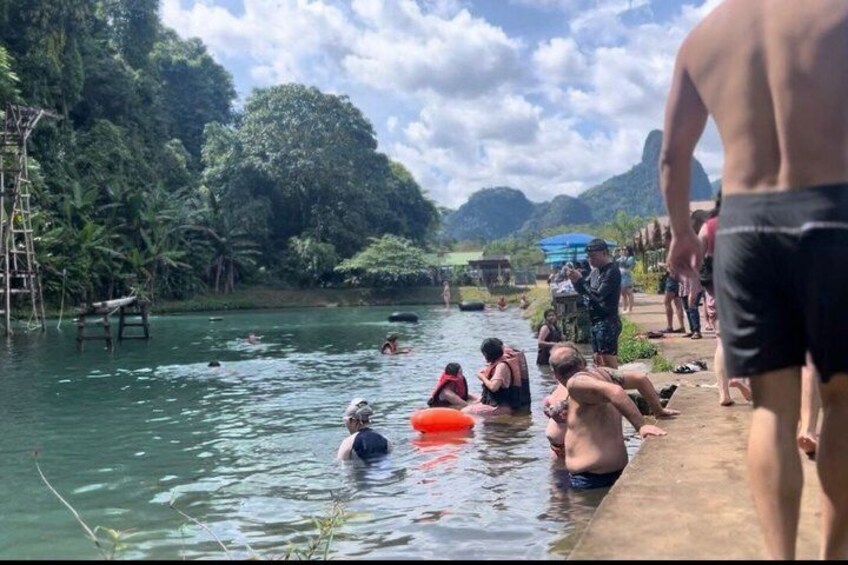 People swimming and sitting by a riverbank in a tropical setting with lush greenery and hills in the background. 4