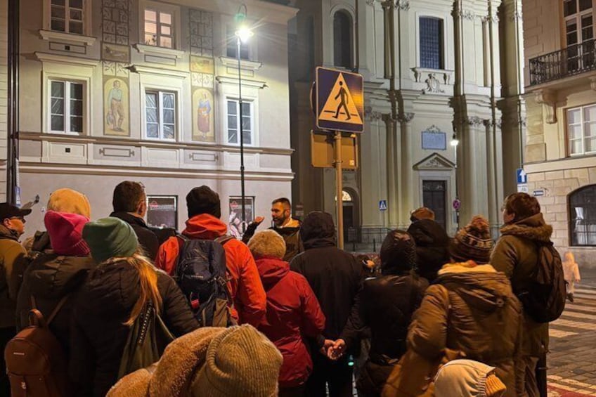 A group of people gathered on a city street at night, with a pedestrian crossing sign and classical architecture in the background. 2