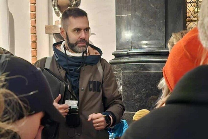 A person in a brown jacket with 'CCTV' text, holding a camera, standing in a public indoor space with marble columns. 3