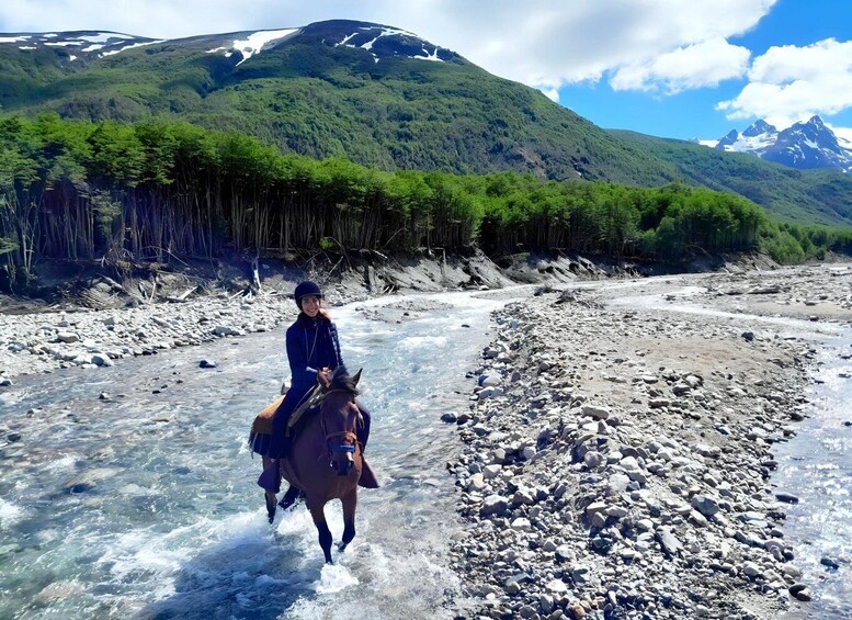 A person riding a horse through a shallow river with a mountainous backdrop. 4