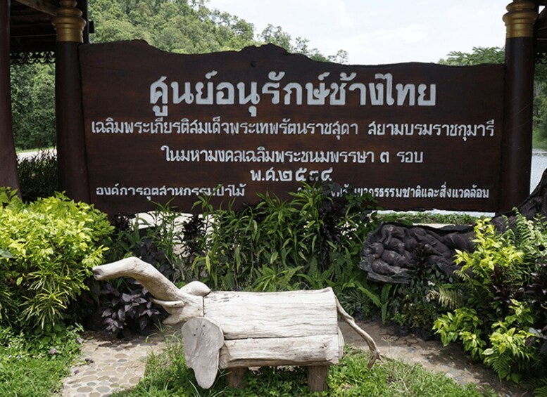 A wooden sign with Thai script, surrounded by greenery and a stone bench. 3