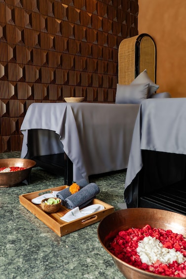 A spa room with two beds, a wooden tray with towels and a bowl of rose petals, and a large bowl of rose petals on the floor. 3