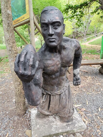 A statue of a muscular figure with a raised fist, set against a backdrop of trees and a sign indicating a 3.6 km trail distance. 3