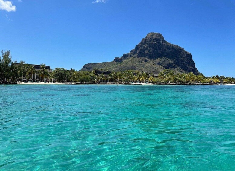 A clear turquoise sea, a mountain, and a beach with palm trees and buildings. 1