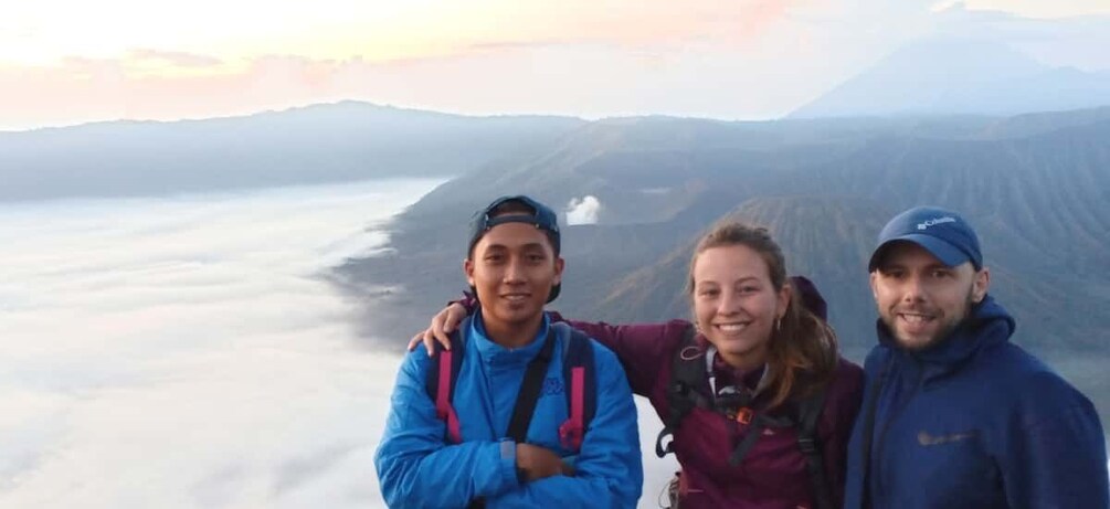 Three people standing with a mountainous landscape in the background. 1