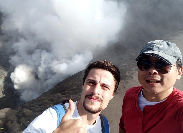 Two people standing in front of a volcanic landscape with smoke rising. 2