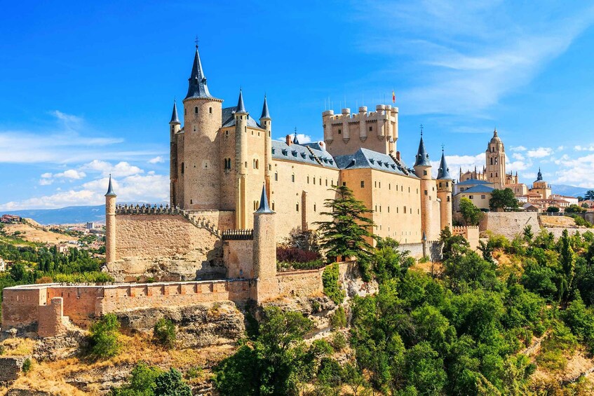 A historic castle with multiple towers and a fortified wall, surrounded by greenery and other buildings. 2